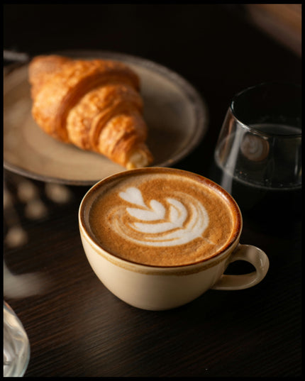 Ein Leinwandbild von einer Tasse Kaffee Latte neben einem Croissant und einem Glas Wasser.