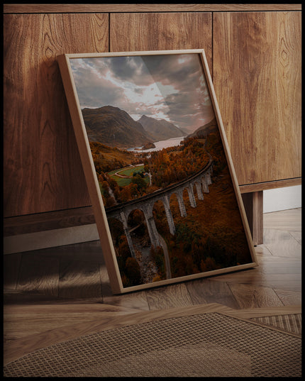 Ein gerahmtes Poster von dem Glenfinnan-Viadukt in Schottland, umgeben von herbstlicher Landschaft und Bergen im Hintergrund, an einer Holzkommode angelehnt und stehend auf dem Holzboden