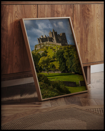 Ein gerahmtes Poster von den Ruinen des Rock of Cashel auf einem Hügel in Irland, umgeben von grünen Wiesen und Bäumen, an einer Holzkommode angelehnt und stehend auf dem Holzboden