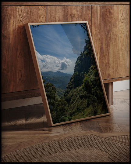 Ein gerahmtes Poster einer bewaldeten Berglandschaft in einer tropischen Region, an einer Holzkommode angelehnt und stehend auf dem Holzboden