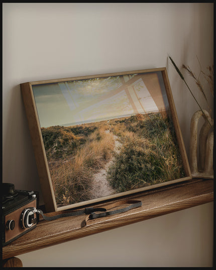 Ein gerahmtes Poster von einer Wanderung durch grasige Dünen, auf einem Holzregal an die Wand angelehnt