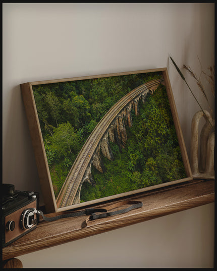 Ein gerahmtes Poster aus der Vogelperspektive von einer Viaduktbrücke aus Stein die durch einen tropischen Wald führt, auf einem Holzregal an die Wand angelehnt