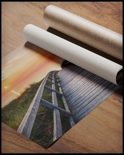 Ein Poster von einem Holzsteg im Sonnenuntergang, der über Dünen zum Strand führt, gerollt auf einer Holzoberfläche liegend