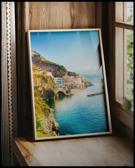 Ein gerahmtes Poster von einer Küstenstadt an der Amalfiküste in Italien, mit Häusern, die an steilen Klippen über dem türkisblauen Meer thronen, vor einem Fenster an die Wand angelehnt