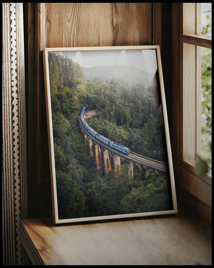 Ein gerahmtes Poster von einem blauen Zug, der über eine alte Brücke inmitten eines grünen Waldes fährt, vor einem Fenster an die Wand angelehnt