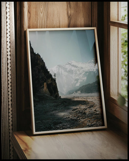 Ein gerahmtes Poster von einem breiten Flussbett in den Dolomiten, umgeben von schneebedeckten Bergen, vor einem Fenster an die Wand angelehnt