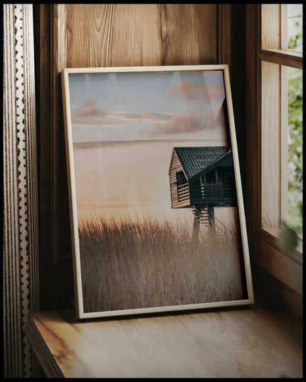 Ein gerahmtes Poster von einer Hütte am Strand in den Dünen bei Sonnenuntergang, vor einem Fenster an die Wand angelehnt