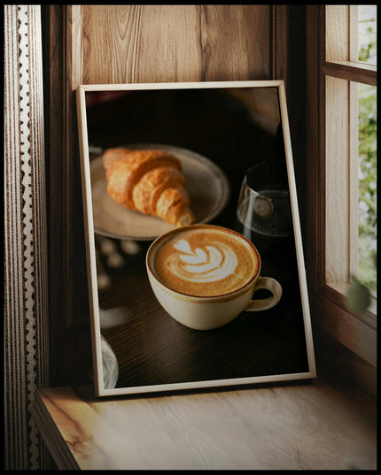 Ein gerahmtes Poster von einer Tasse Kaffee Latte neben einem Croissant und einem Glas Wasser, vor einem Fenster an die Wand angelehnt