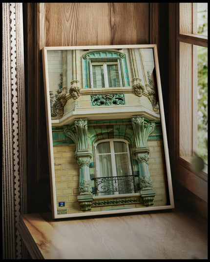 Ein gerahmtes Poster von einer grünlich, beigen Häuserfassade aus Stein mit Balkon und Verzierungen, vor einem Fenster an die Wand angelehnt