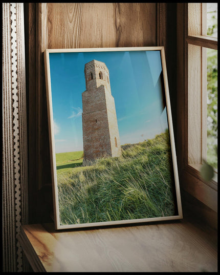 Ein gerahmtes Poster von einem steinernen Turm auf einer grünen, hügeligen Wiese, vor einem Fenster an die Wand angelehnt