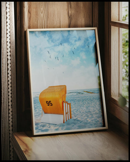 Ein gerahmtes Poster von einem orangenen Strandkorb am Strand der in Richtung Meer gerichtet ist, vor einem Fenster an die Wand angelehnt